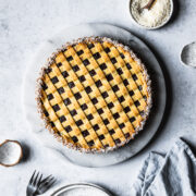 A baked pasta frola (quince tart) with thin lattice on top and a border of dessicated coconut. The tart rests on a marble platter on a blue-grey stone surface and is surrounded by small bowls and a blue linen napkin.