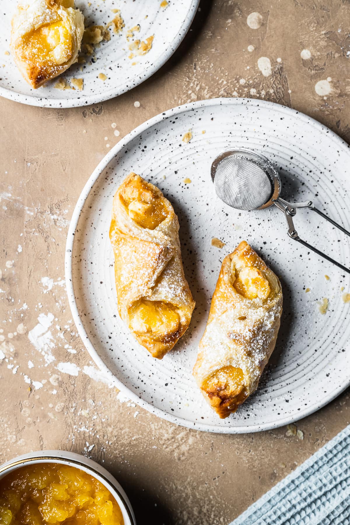 Two golden brown filled quesito pastries on a speckled white ceramic plate. A small metal sieve of powdered sugar rests on the right of the plate. A small bowl of pineapple jam and a light blue napkin peek into the frame at bottom. Another plate peeks into the frame at top left. The plates rest on a tan stone surface.