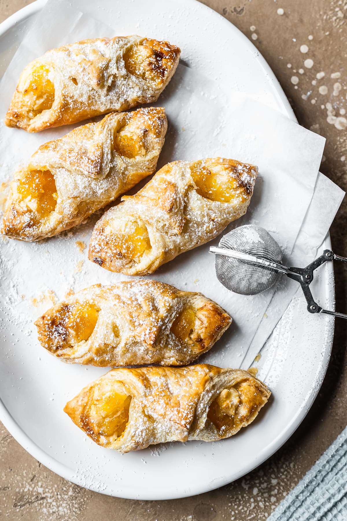 Five golden brown filled Puerto Rican quesitos on white parchment on a white ceramic plate. A small metal sieve of powdered sugar rests on the right of the plate. A bit of a light blue napkin peeks into the frame at bottom right. The plate rests on a tan stone surface.