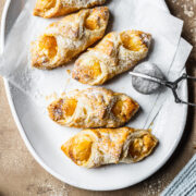 Baked pastries on white parchment on a white ceramic plate. A small metal sieve of powdered sugar rests on the right of the plate. A bit of a light blue napkin peeks into the frame at bottom right. The plate rests on a tan stone surface.