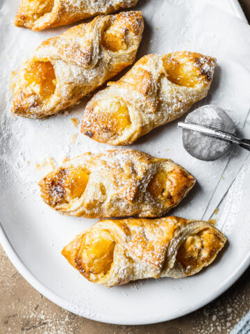 Baked pastries on white parchment on a white ceramic plate. A small metal sieve of powdered sugar rests on the right of the plate. A bit of a light blue napkin peeks into the frame at bottom right. The plate rests on a tan stone surface.