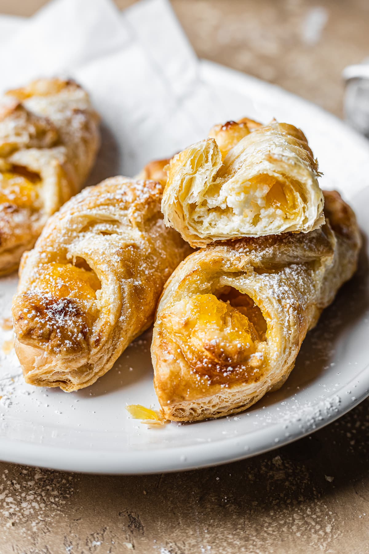 A close up image of cream cheese and pineapple jam pastries on a white ceramic plate ona warm tan stone surface. One of the quesitos has been cut open to show the flaky layers and filling.