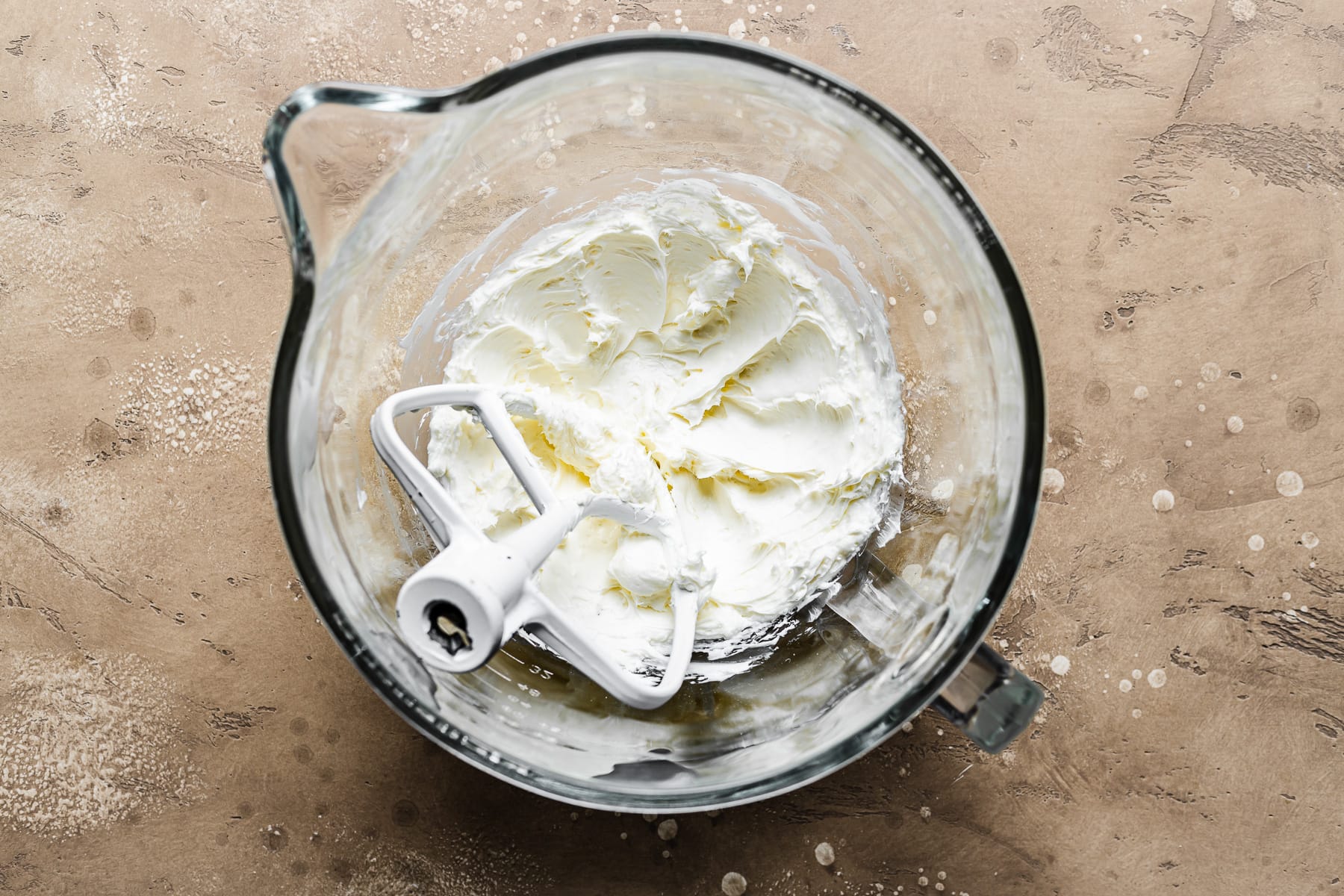 A process photo of cream cheese filled pastries showing a glass mixing bowl with cream cheese and sugar blended together. The bowl rests on a warm tan stone surface.