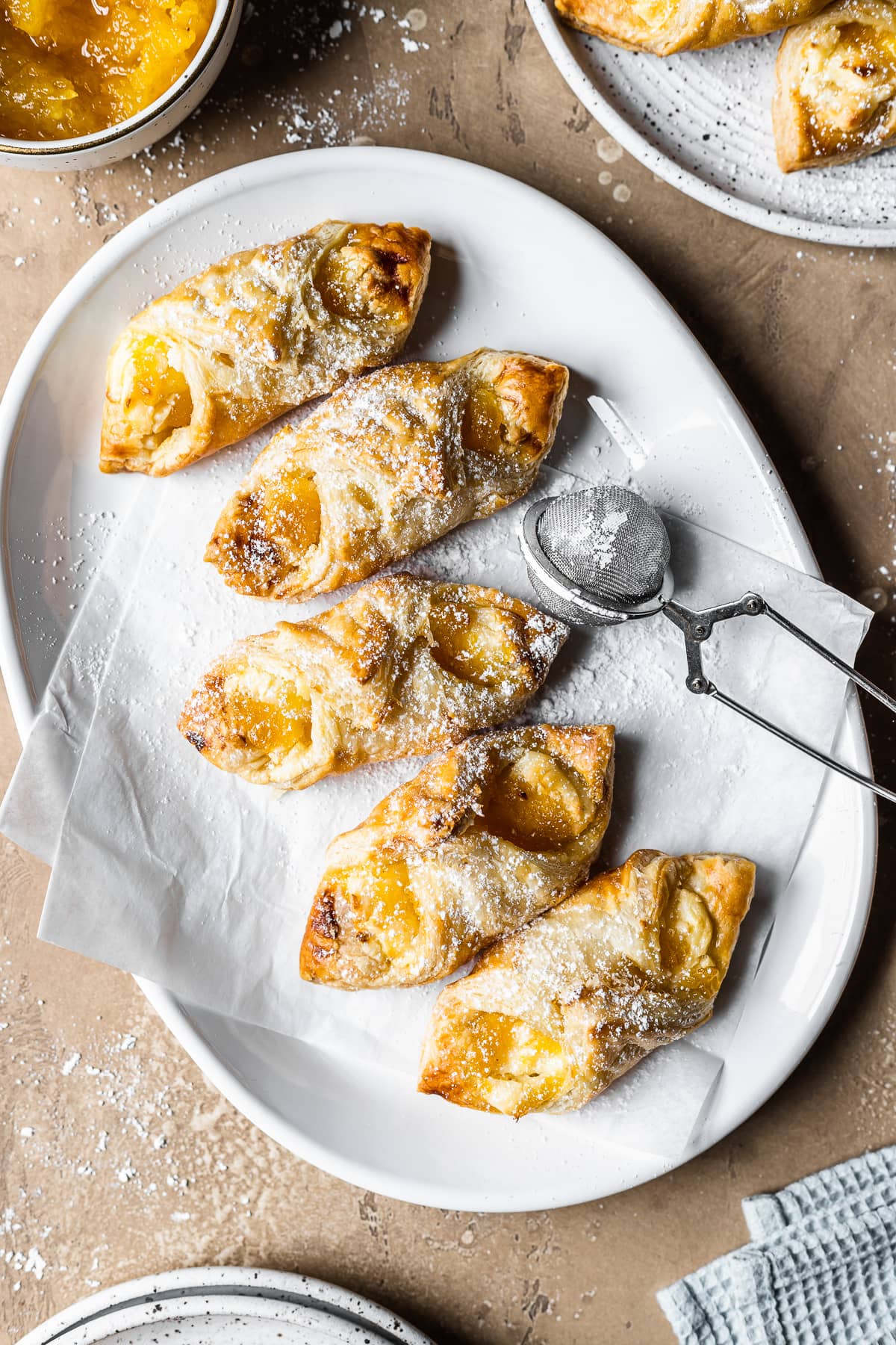 Five golden brown Puerto Rican quesitos on white parchment on a white ceramic plate on a warm tan stone surface. A small metal sieve of powdered sugar rests on the right of the plate. A bit of a light blue napkin peeks into the frame at bottom right. A bowl of jam and a plate of additional pastries peek into the frame at top.