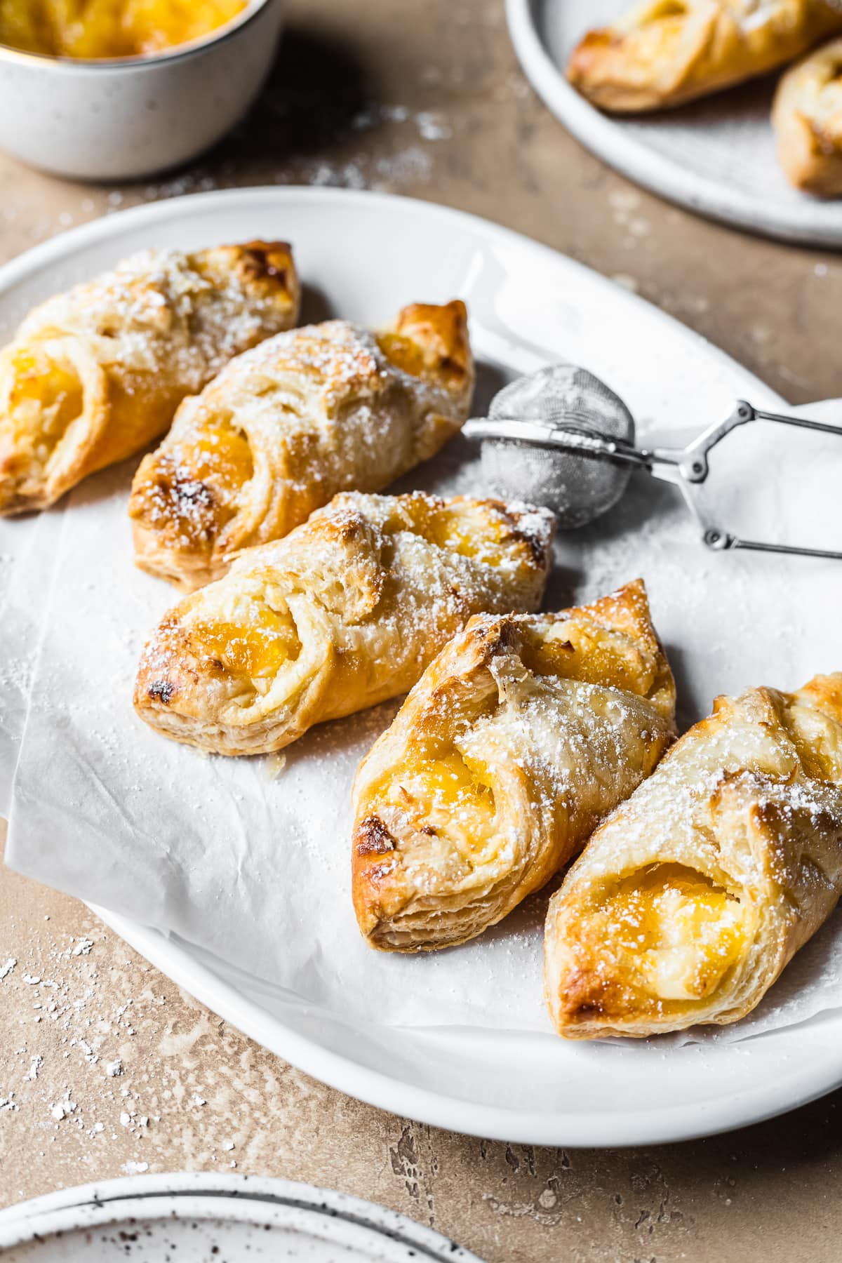 Five golden brown quesitos on white parchment on a white ceramic plate on a tan stone surface. A small metal sieve of powdered sugar rests on the right of the plate. A small bowl of pineapple jam and a small plate with more pastries peek into the frame at top.
