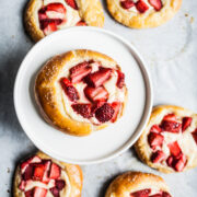 Brioche pastries with strawberries and cream cheese on white parchment paper. One pastry is elevated on a white ceramic plate.