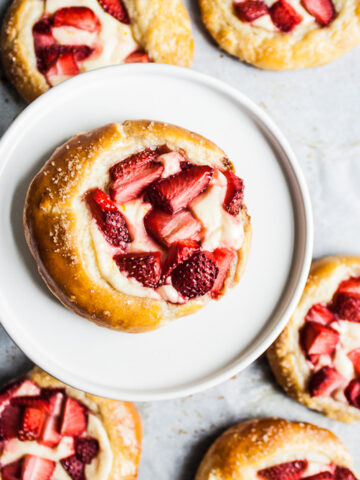 Brioche pastries with strawberries and cream cheese on white parchment paper. One pastry is elevated on a white ceramic plate.