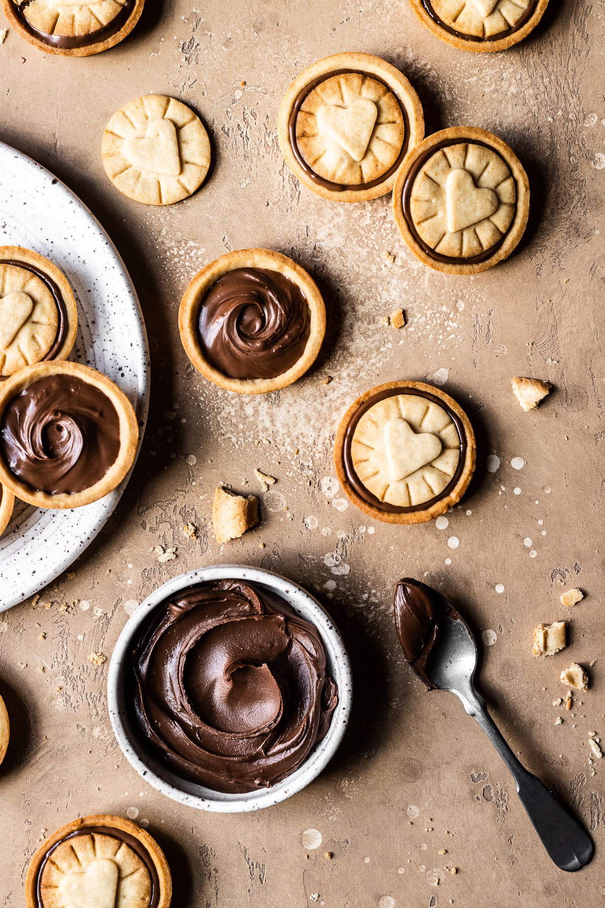 Chocolate hazelnut filled cookies on a warm tan stone surface. A small white ceramic bowl of Nutella rests at bottom with a silver spoon nearby. A white ceramic plate full of cookies peeks into the photo at left.