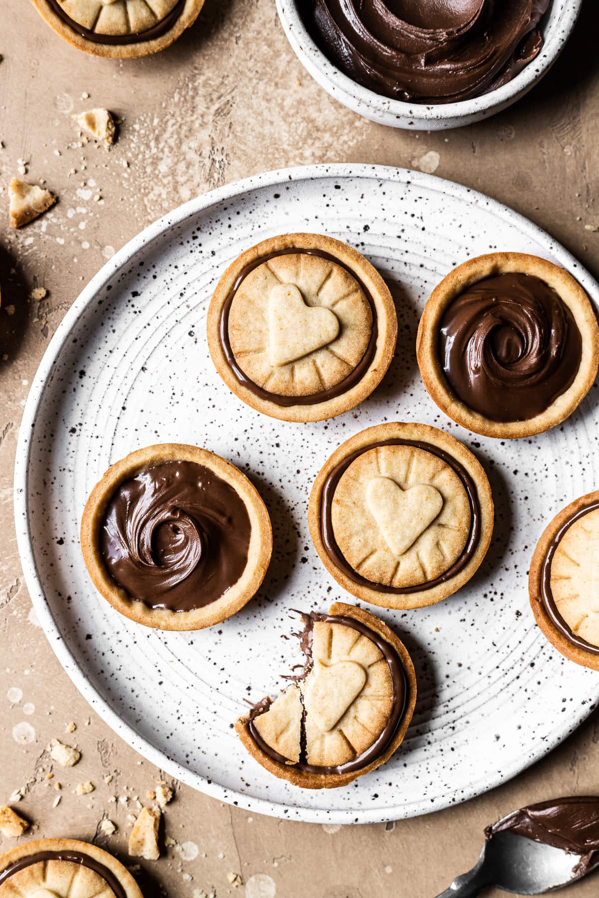 Golden brown honey Nutella biscuits on a white speckled ceramic plate. A small bowl of Nutella rests nearby. The plate sits on a warm tan stone surface. More cookies and crumbs surround the plate.