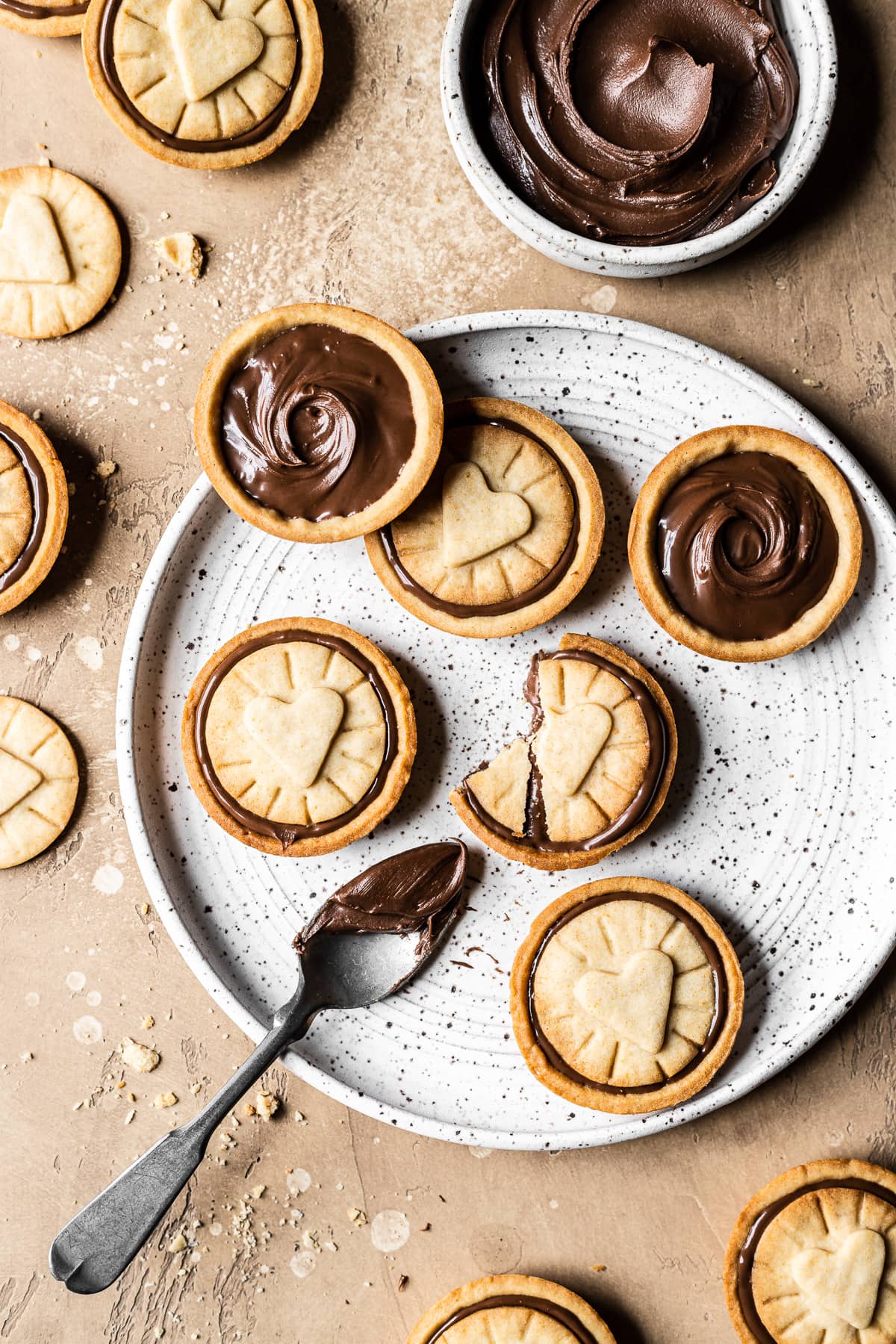 Nutella filled biscuits on a white speckled ceramic plate. A small bowl of Nutella rests nearby. A spoon of Nutella rests on the plate. The plate sits on a warm tan stone surface. More cookies and crumbs surround the plate.