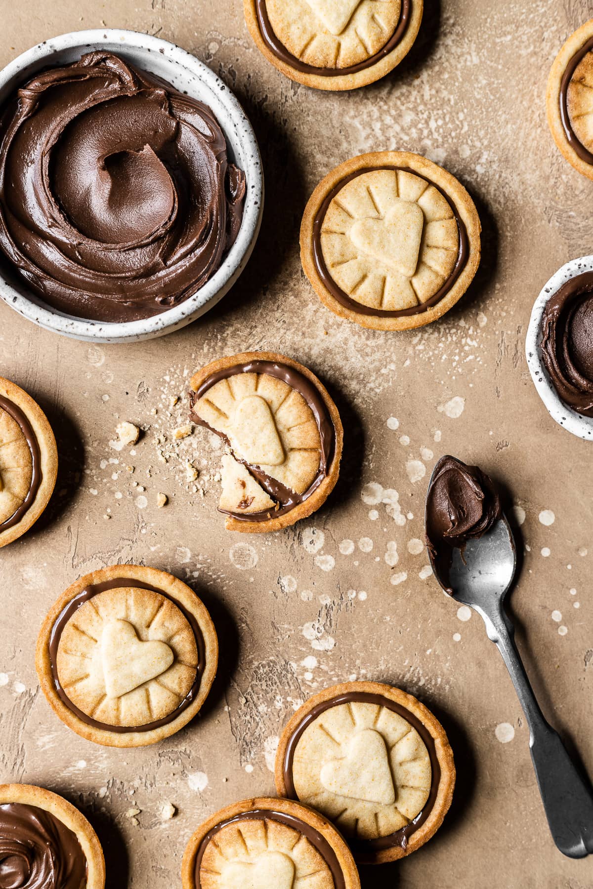 Nutella stuffed cookies on a warm tan speckled stone surface. A small white ceramic bowl is filled with a swirl of Nutella. An antique silver spoon with a scoop of Nutella rests on the surface at bottom right.