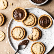 Nutella filled biscuits on a white speckled ceramic plate. A small bowl of Nutella rests nearby. A spoon of Nutella rests on the plate. The plate sits on a warm tan stone surface. More cookies and crumbs surround the plate.