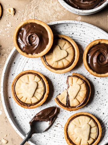 Nutella filled biscuits on a white speckled ceramic plate. A small bowl of Nutella rests nearby. A spoon of Nutella rests on the plate. The plate sits on a warm tan stone surface. More cookies and crumbs surround the plate.