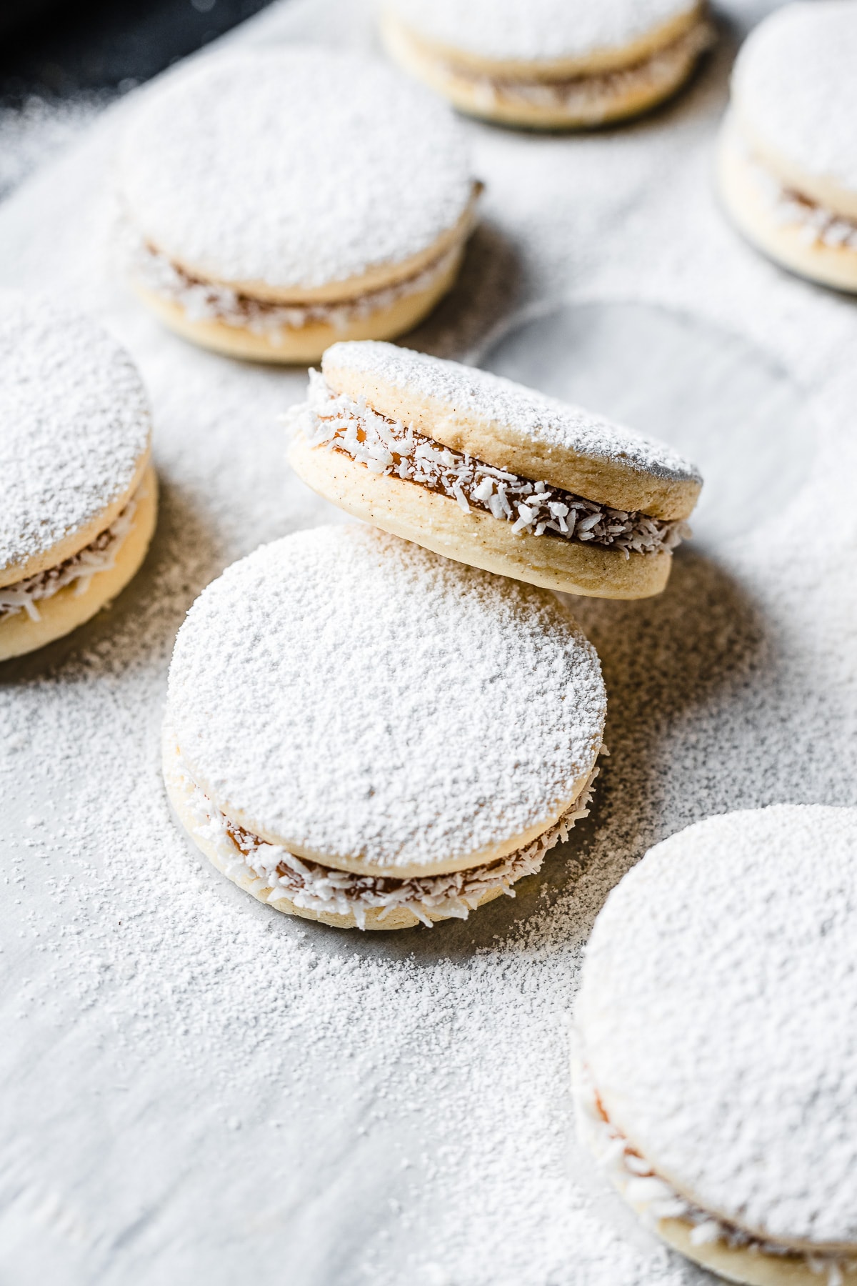 A close up photo showing Argentine shortbread cookies dusted with powdered sugar on a parchment lined cookie sheet. One cookie rests on another at an angle to show the filling.