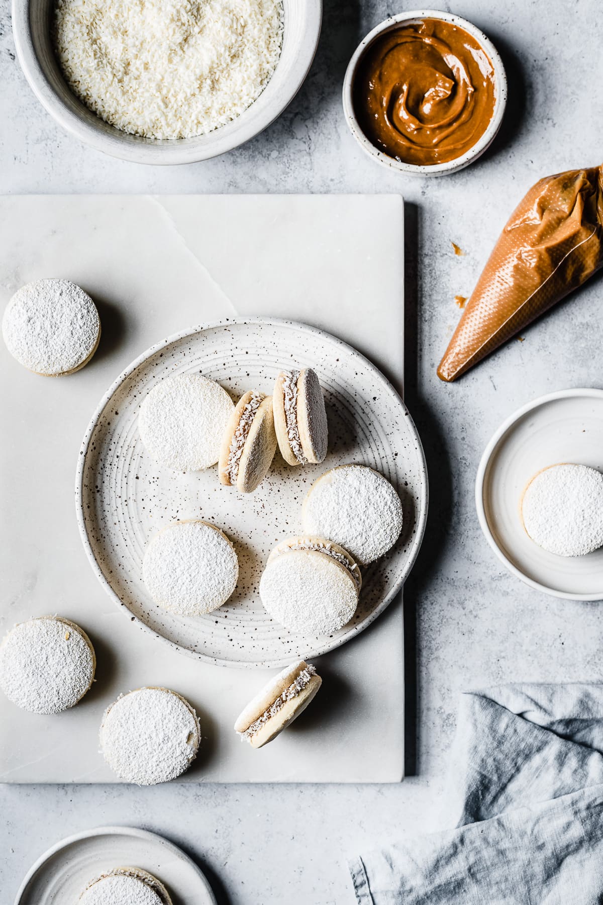 Flatlay image of filled Argentine shortbread cookies on a white plate on a marble platter on a light cement surface. Shredded coconut, dulce de leche, more cookies and a light blue linen napkin surround the scene.
