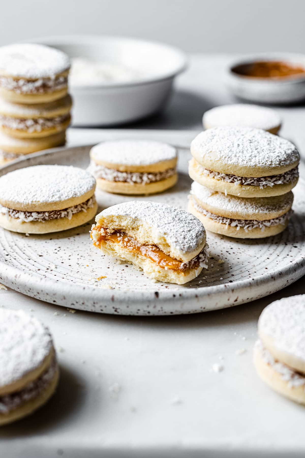 A light and airy image of alfajores de maicena on a white speckled ceramic plate. One cookie is partially eaten, showing the dulce de leche inside. More cookies are nearby, with a bowls of powdered sugar and dulce de leche in the background.