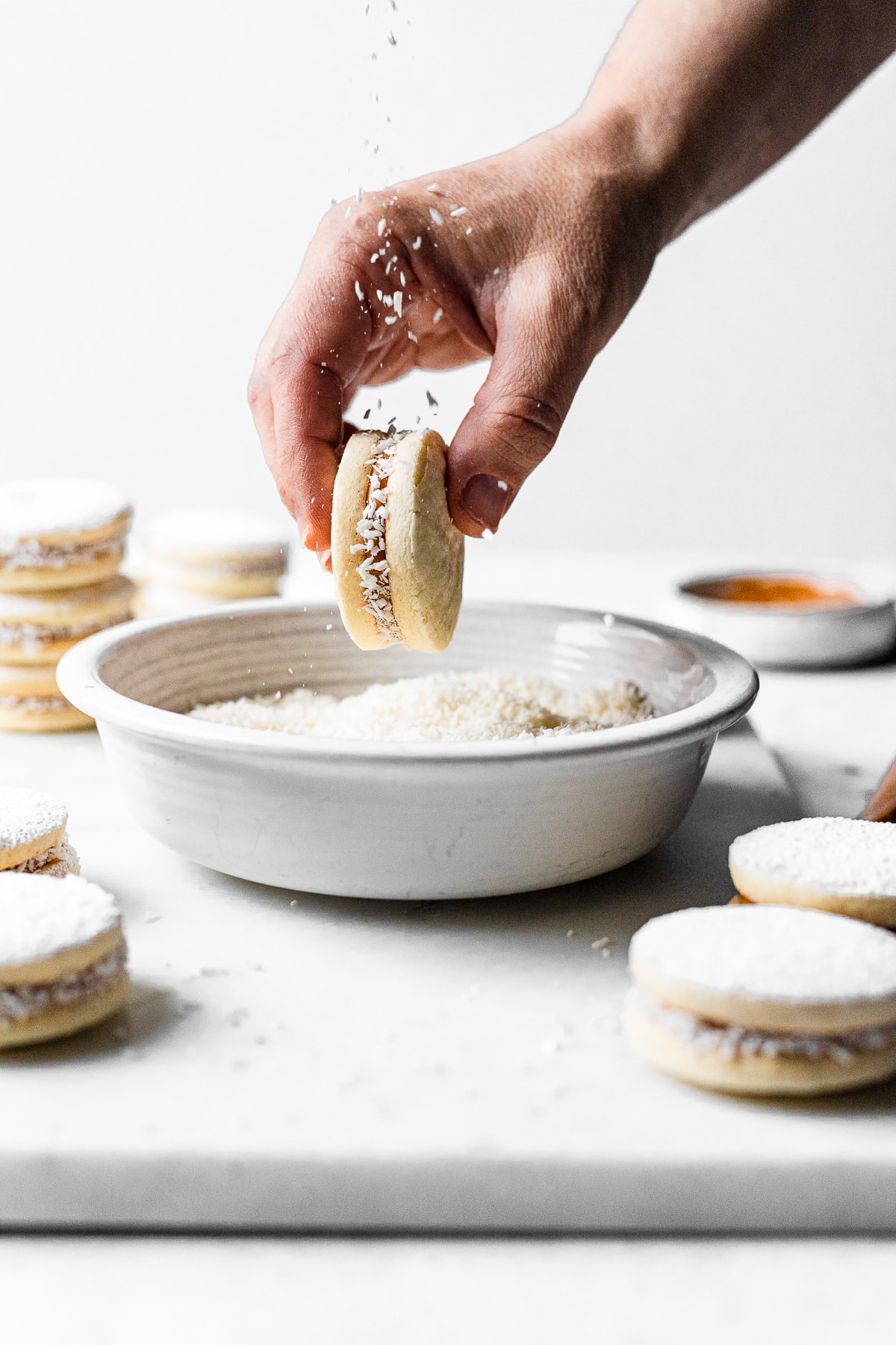 A light and airy image of a hand holding an alfajor cookie over a bowl of shredded coconut. More cookies are nearby. There is a sprinkling of shredded coconut being dropped from above.