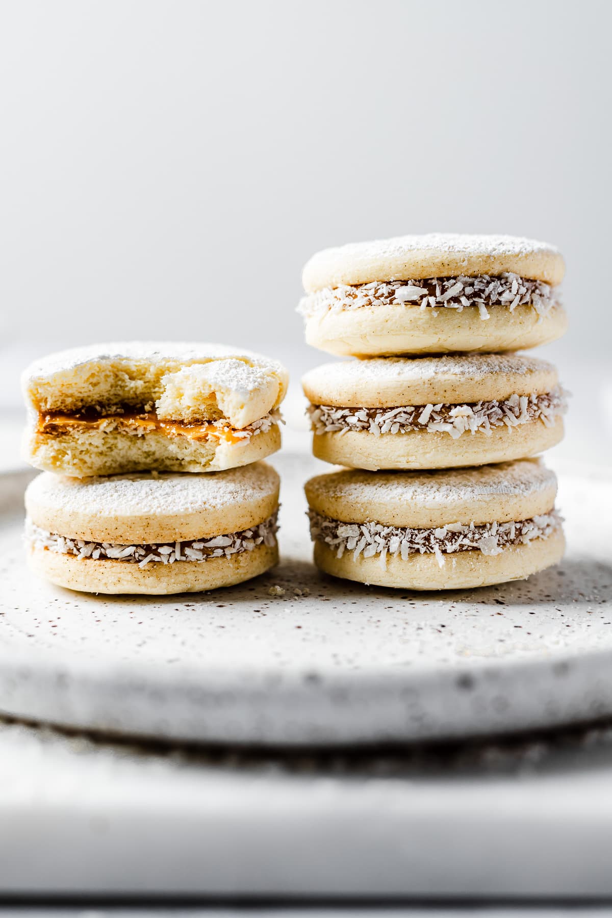 Two stacks of Argentine alfajores de maicena. The stack on the left has two cookies. The top one has a bite taken out of it.