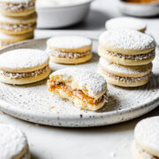 A light and airy image of alfajores de maicena on a white speckled ceramic plate. One cookie is partially eaten, showing the dulce de leche inside. More cookies are nearby, with small white bowls in the background.