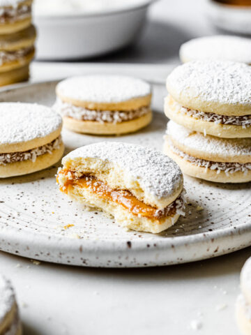 A light and airy image of alfajores de maicena on a white speckled ceramic plate. One cookie is partially eaten, showing the dulce de leche inside. More cookies are nearby, with small white bowls in the background.