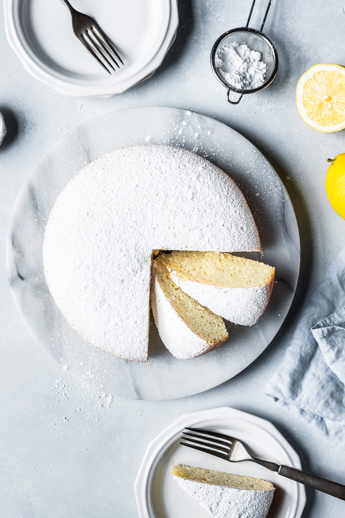 Top view of a pale yellow light and fluffy torta paradiso (paradise cake) covered in powdered sugar on a round marble platter. Two slices are turned at an angle, showing the fine pale yellow texture. A pale blue linen napkin rests nearby along with lemons.