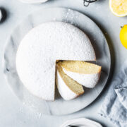Top view of a pale yellow light and fluffy torta paradiso (paradise cake) covered in powdered sugar on a round marble platter. Two slices are turned at an angle, showing the fine pale yellow texture. A pale blue linen napkin rests nearby along with lemons.