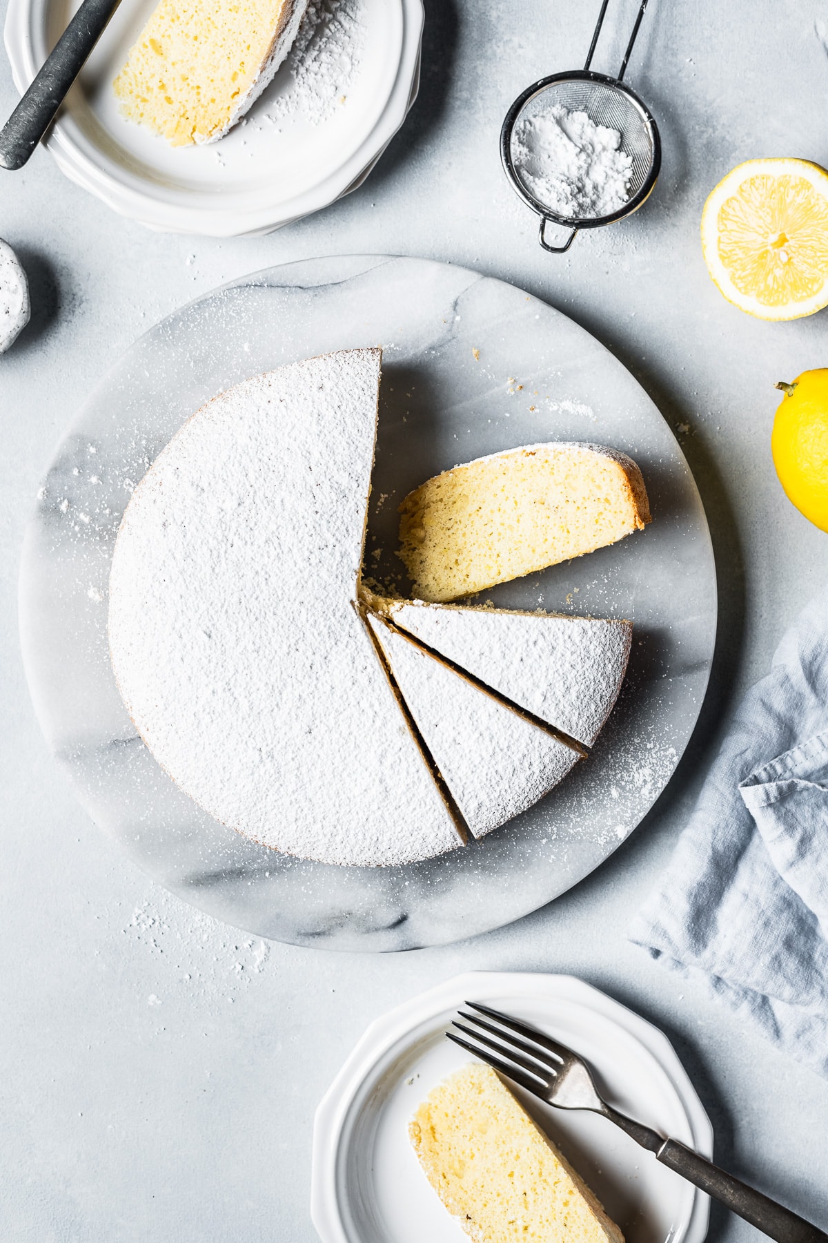 Top view of a light and fluffy cake covered in powdered sugar on a round marble platter. Several slices have been cut and a few rest on nearby white plates. One slice is turned on its side, showing the fine pale yellow texture. A pale blue linen napkin rests nearby along with lemons and a sieve of powdered sugar.