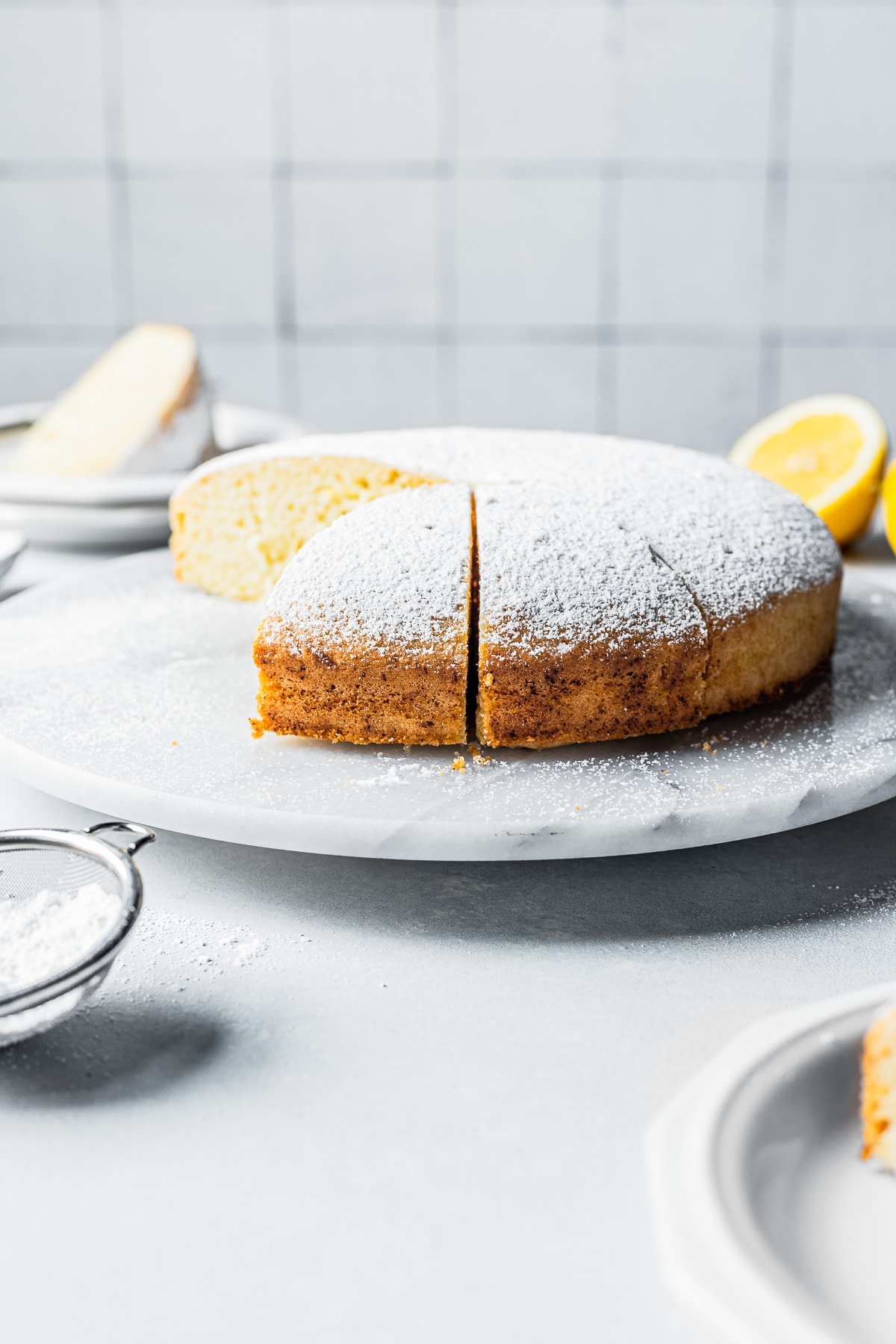 Side view of torta paradiso (paradise cake) topped with powdered sugar. The cake is on a grey marble platter on a light blue surface. There is a rustic tile background. Plates with slices of cake and lemons are in the background.