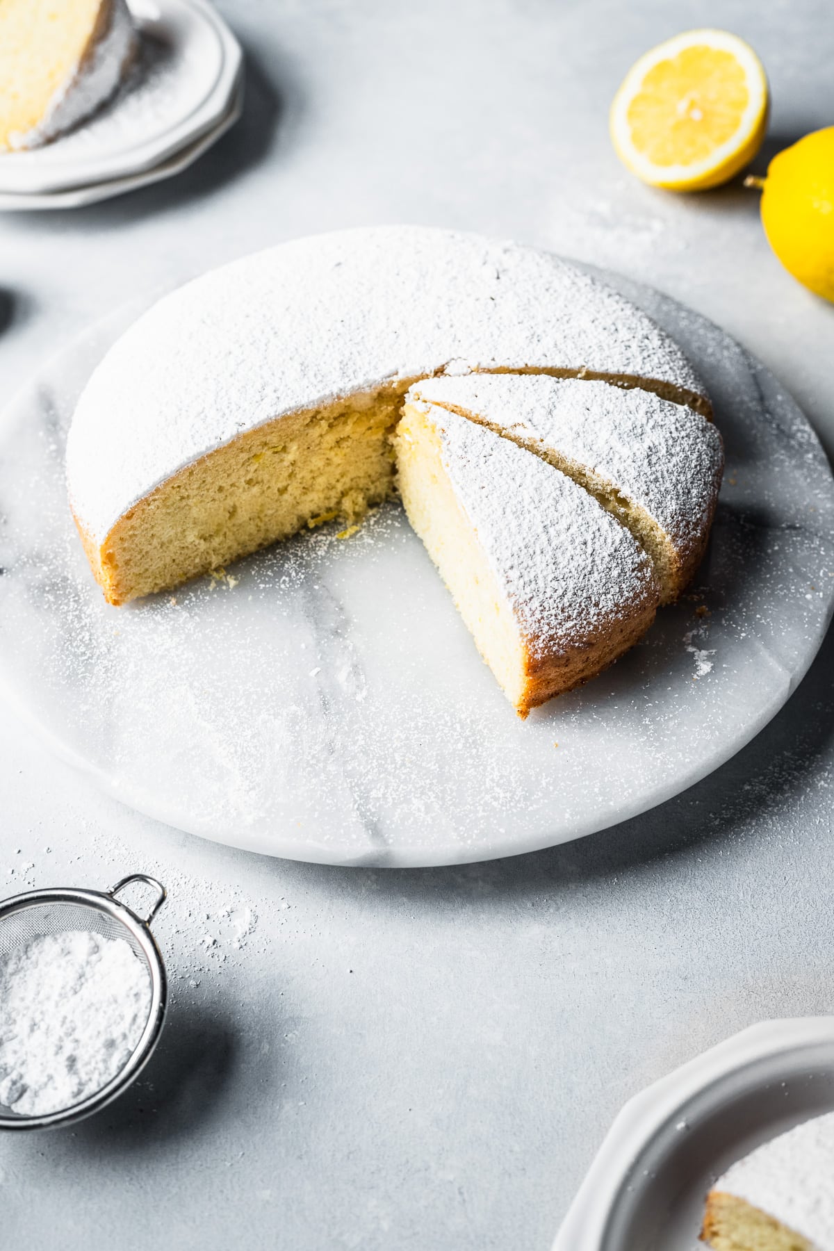 Slices of torta paradiso cake on a round white marble platter on a light blue surface. Some slices are nearby on white ceramic plates, revealing the center crumb of the cake.