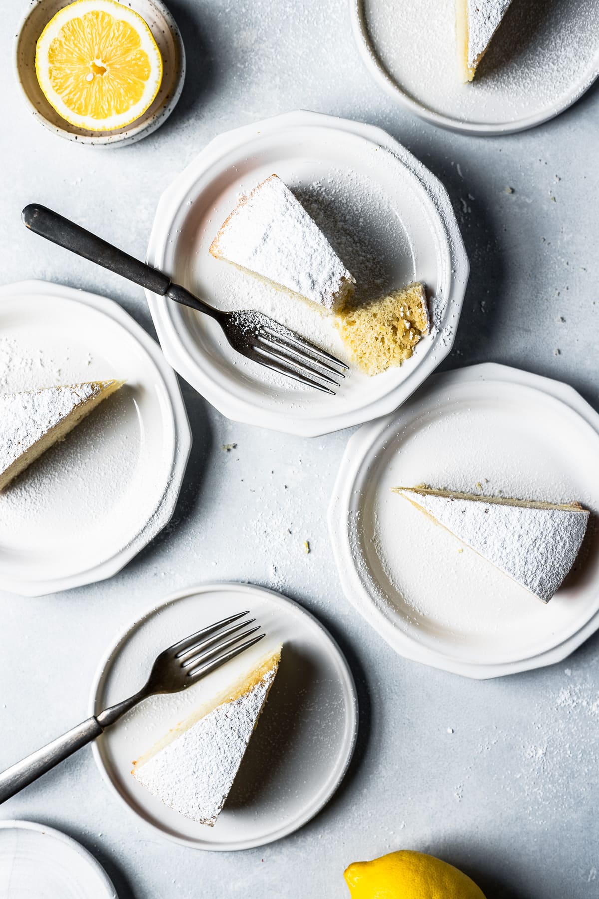 5 slices of torta paradiso (paradise cake) on white ceramic plates on a light blue surface. The slices have been dusted with powdered sugar. A bite has been taken from one piece. Lemons rest at the top and bottom of the frame.