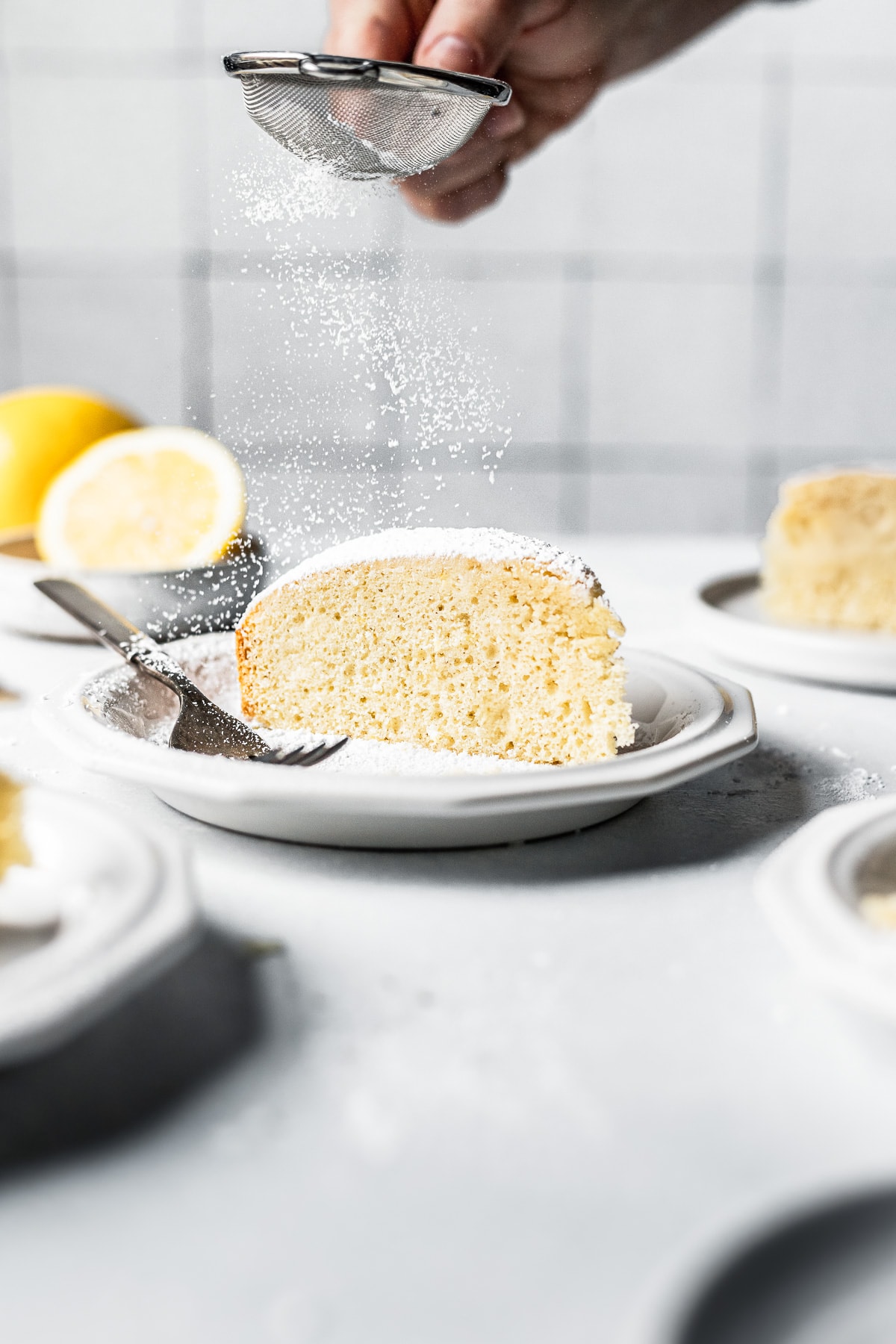 A hand sprinkles a small sieve of powdered sugar over the top of a slice of light and airy paradise cake. The plate is surrounded by other plates and a small bowl of lemons.