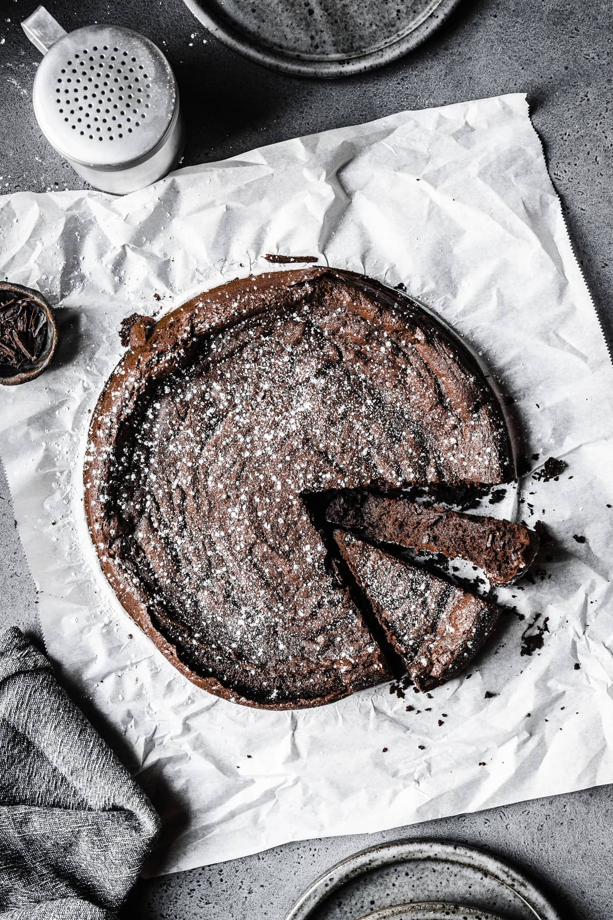 A dense dark chocolate cake sprinkled with powdered sugar on a piece of white parchment paper. Two slices are cut into the cake, one turned on its side to show the interior. The parchment paper rests on a grey surface.