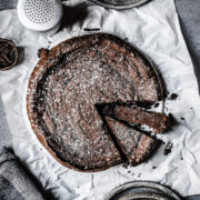 A dense dark chocolate cake sprinkled with powdered sugar on a piece of white parchment paper. Two slices are cut into the cake, one turned on its side to show the interior. The parchment paper rests on a grey surface.