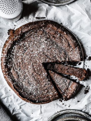 A dense dark chocolate cake sprinkled with powdered sugar on a piece of white parchment paper. Two slices are cut into the cake, one turned on its side to show the interior. The parchment paper rests on a grey surface.