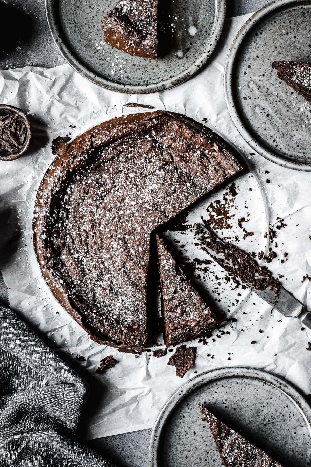 A flourless dark chocolate torta tenerina with slices cut out and placed on grey ceramic plates. A crumb covered knife rests near the rest of the cake, and a grey napkin is at bottom left. There are crumbs scattered around the cake.