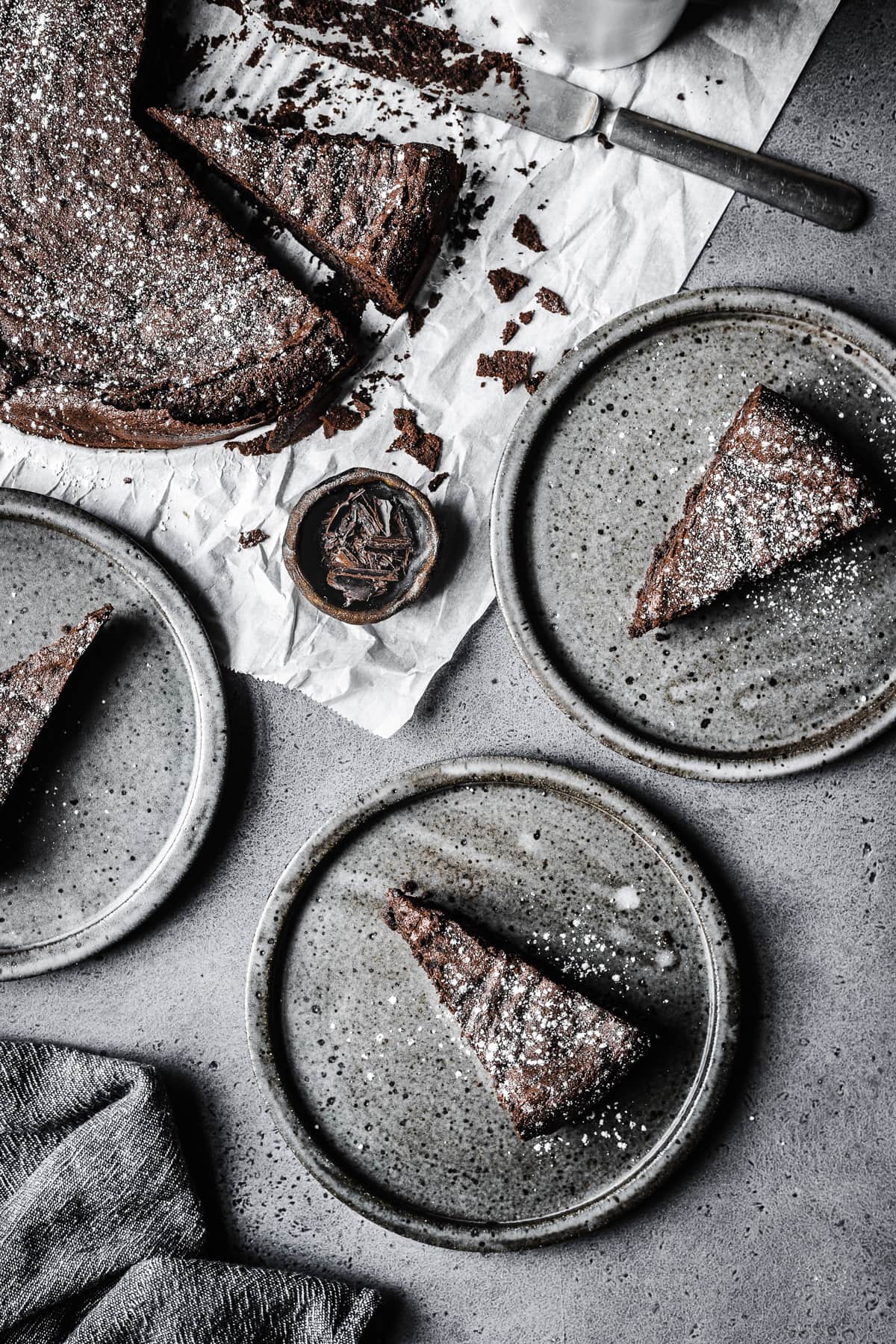 Slices of flourless chocolate cake on grey ceramic plates on a grey stone surface. The remainder of the cake rests on a piece of white parchment paper along with a vintage knife covered in crumbs.