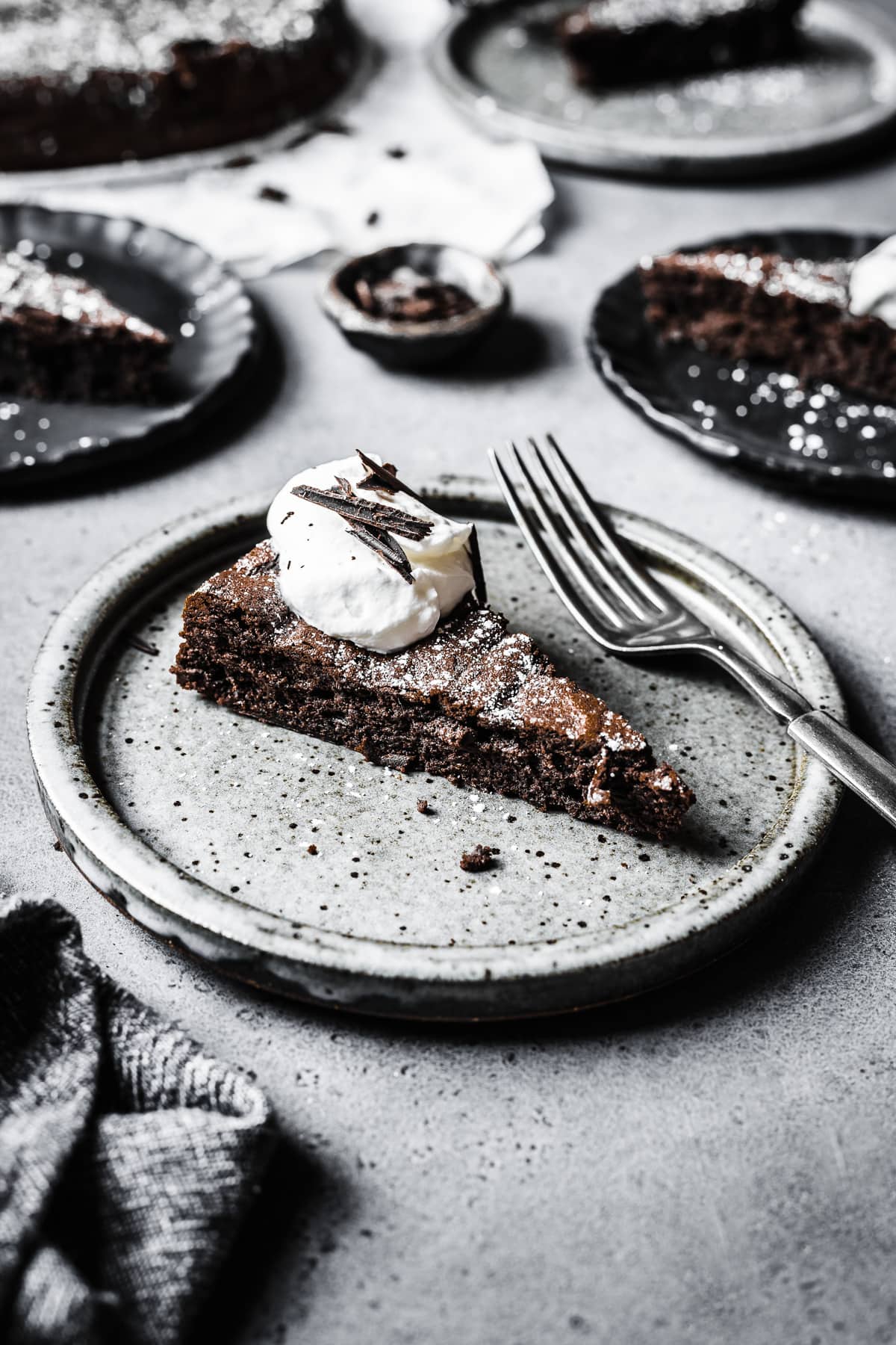 A slice of flourless torta tenerina with a bit of whipped cream and chocolate shavings on top on a grey ceramic plate resting on a grey stone surface. There are more slices on ceramic plates in the background, along with the rest of the cake.