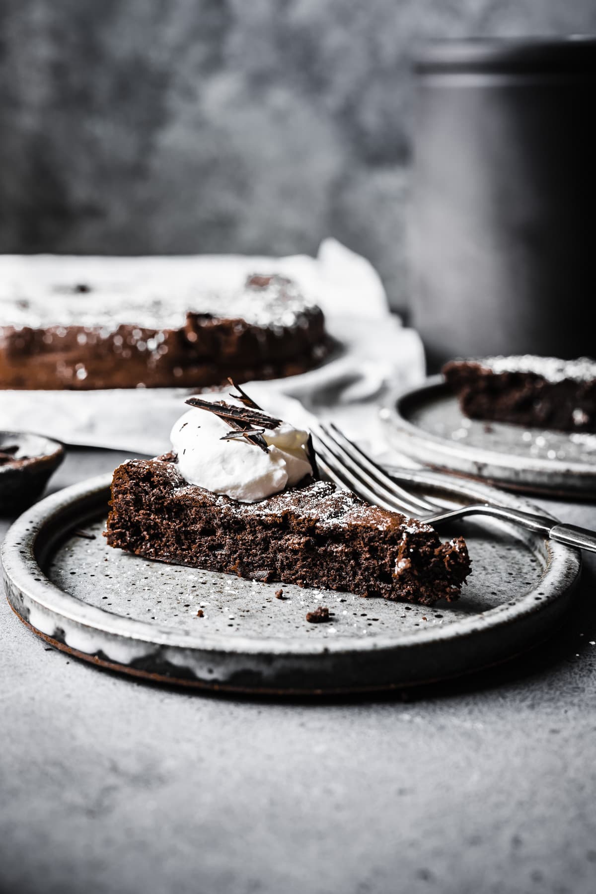 A slice of flourless chocolate cake with whipped cream and chocolate shavings on a ceramic plate, viewed from the side to show the cake's crumb. The remainder of the cake is out of focus in the background.