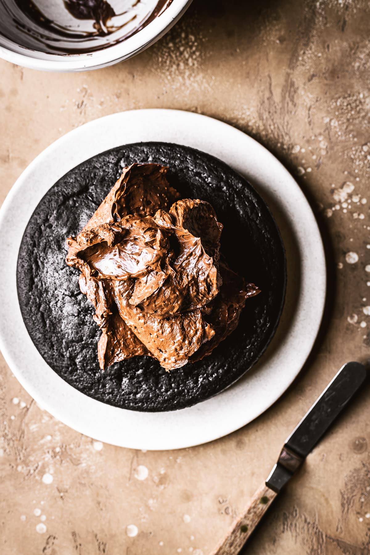 A single layer of dark chocolate cake on a tan plate with a pile of chocolate ganache waiting to be spread. A mini offset spatula rests nearby. The plate rests on a warm speckled tan surface.