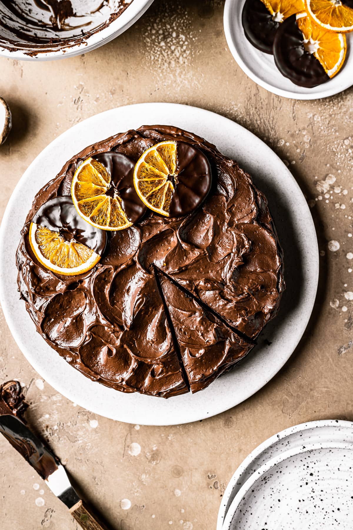 An orange chocolate cake with a slice cut but not removed. The ganache frosting is decorated with rustic swirls and three dried oranges dipped in chocolate. The cake is on a tan plate on a warm brown stone background.