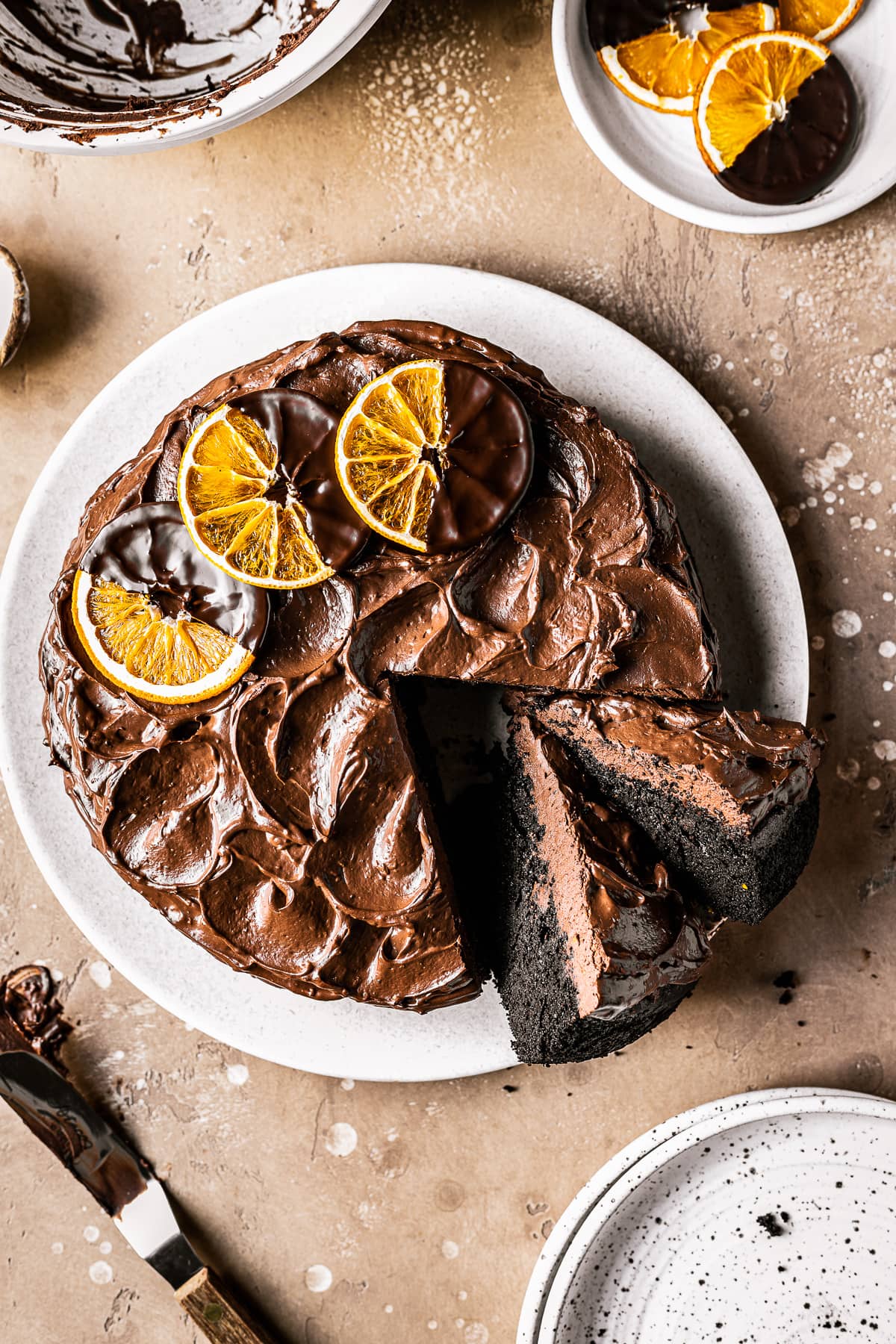 A chocolate cake with two slices cut out and tipped to the side revealing the dark interior and fudgy chocolate frosting. The frosting is decorated with rustic swirls and three dried oranges dipped in chocolate. The cake is on a tan plate on a warm brown stone background.