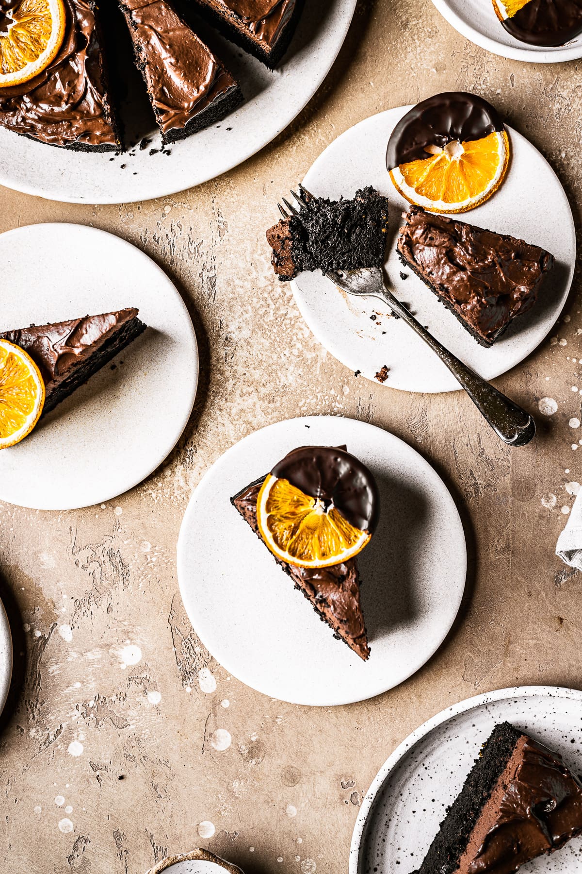 Top view of slices of chocolate orange cake on tan plates on a warm tan stone surface. The cake slices have chocolate frosting on top and are topped with dried orange slices dipped in chocolate.