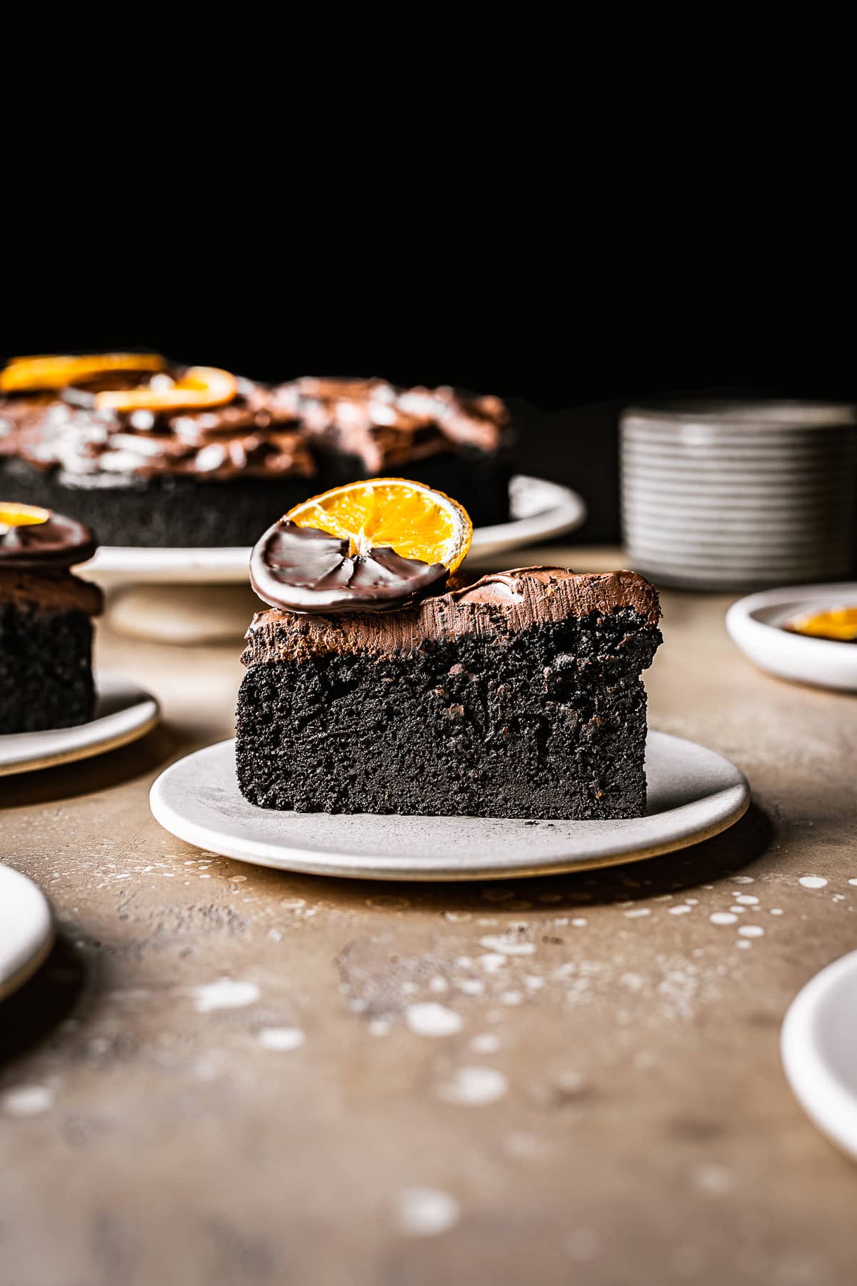 Slice of chocolate orange cake on a plate with the remainder of the cake in the background. Plate is resting on a speckled warm tan stone surface.