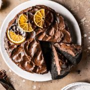 A chocolate cake with two slices cut out and tipped to the side revealing the dark interior and fudgy chocolate frosting. The frosting is decorated with rustic swirls and three dried oranges dipped in chocolate. The cake is on a tan plate on a warm brown stone background.