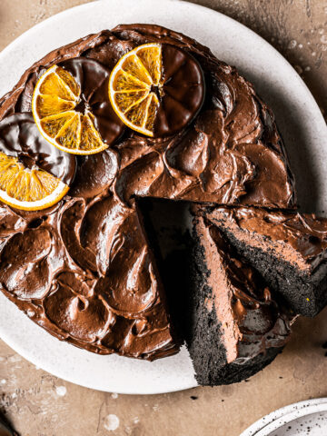 A chocolate cake with two slices cut out and tipped to the side revealing the dark interior and fudgy chocolate frosting. The frosting is decorated with rustic swirls and three dried oranges dipped in chocolate. The cake is on a tan plate on a warm brown stone background.