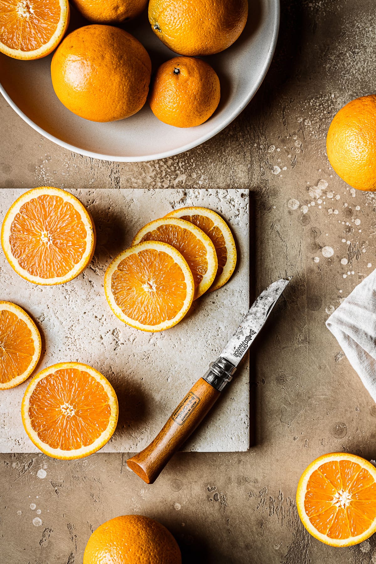 Thinly sliced fresh oranges on a stone cutting surface with a knife resting nearby. The background is a warm stone speckled tan color. A large bowl of oranges rests at top left.