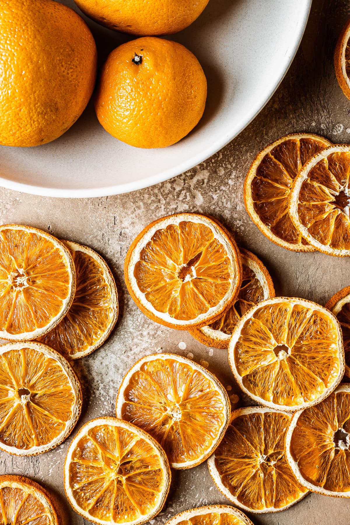 Dried orange slices on a warm tan background with a white bowl of oranges of different sizes at top left.
