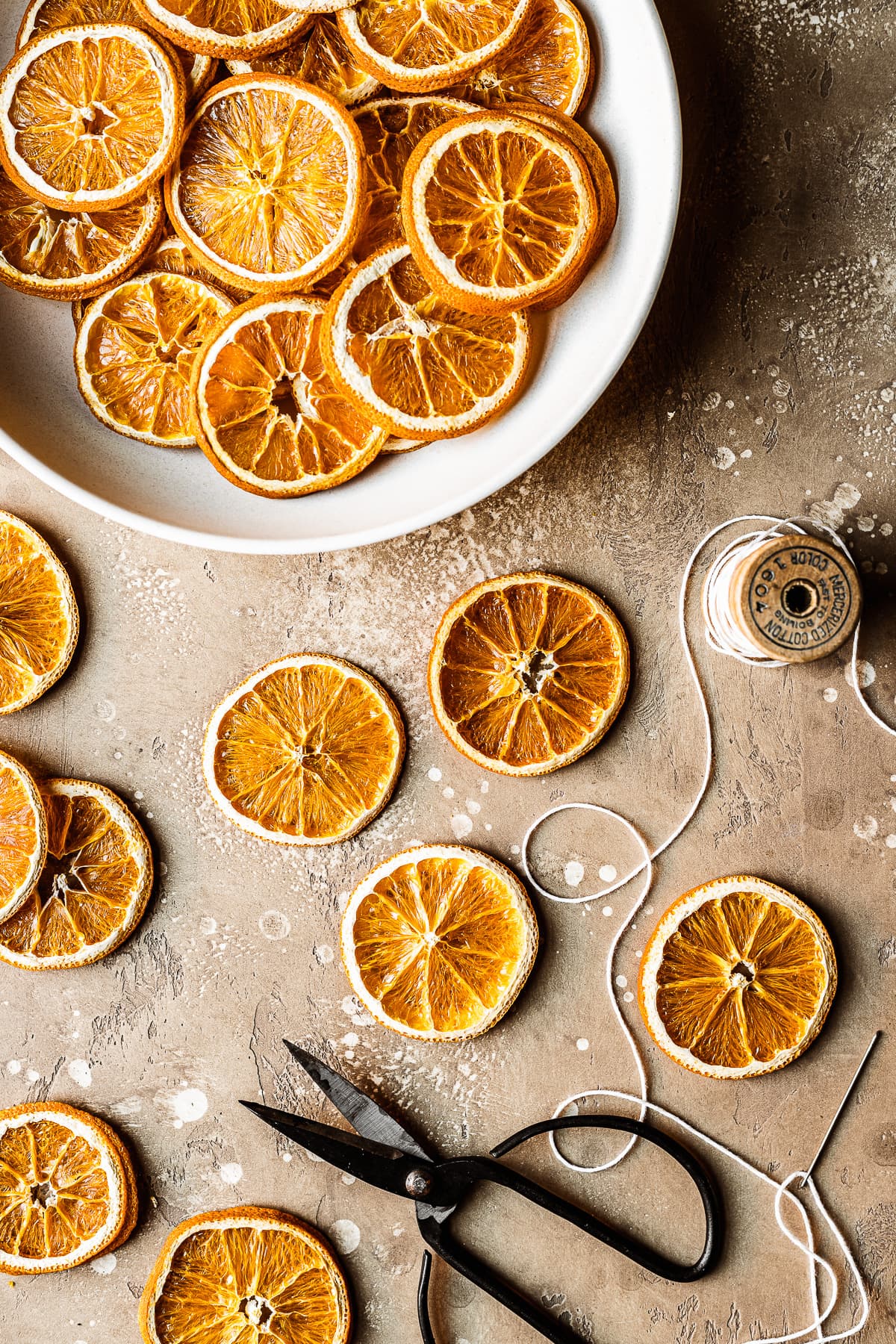 Dried orange slices in a white bowl and resting on a tan speckled surface. A pair of scissors and a spool of thread with a needle rest nearby to make the citrus slices into a garland.