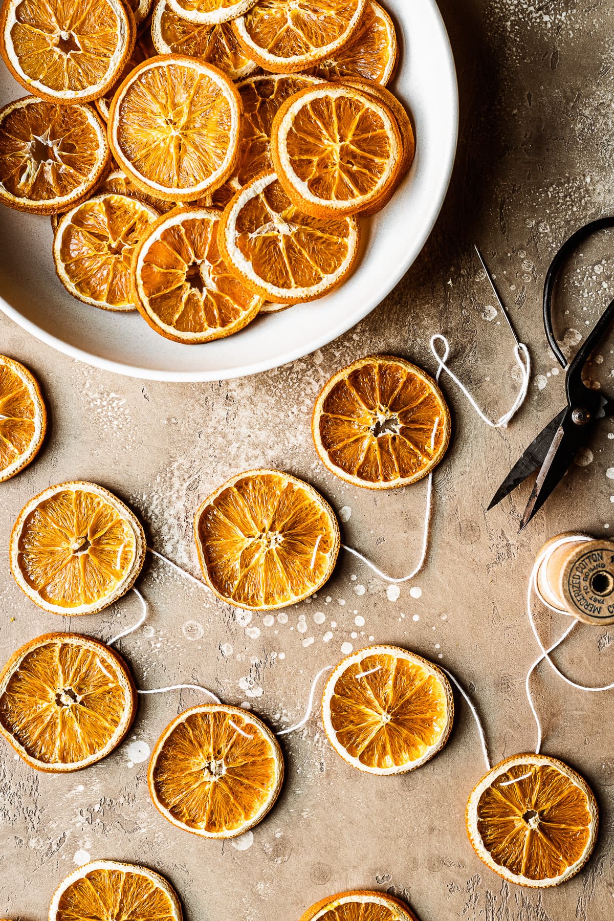 A garland of dried orange slices on a speckled tan surface. There is a large bowl of additional orange slices at top left.