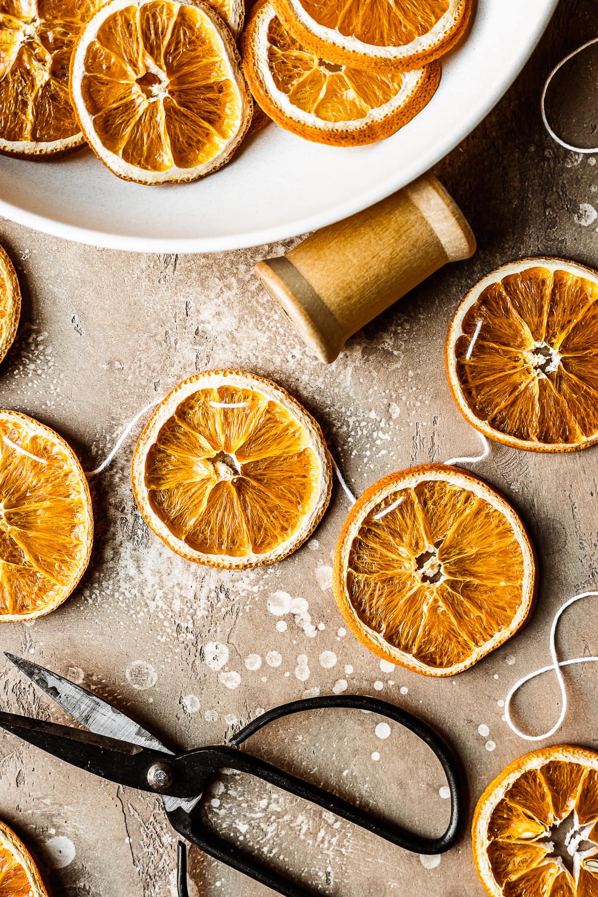 A close up photo of a dried citrus garland with a pair of black scissors nearby. A low white bowl of dried citrus rests nearby at top left near a wooden spool.